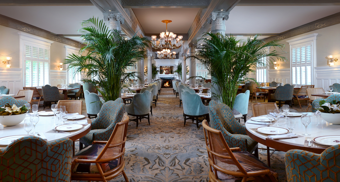 Elegant restaurant dining room with round tables, upholstered chairs, large potted plants, chandeliers, and a fireplace at the far end. Natural light streams in from multiple windows.
