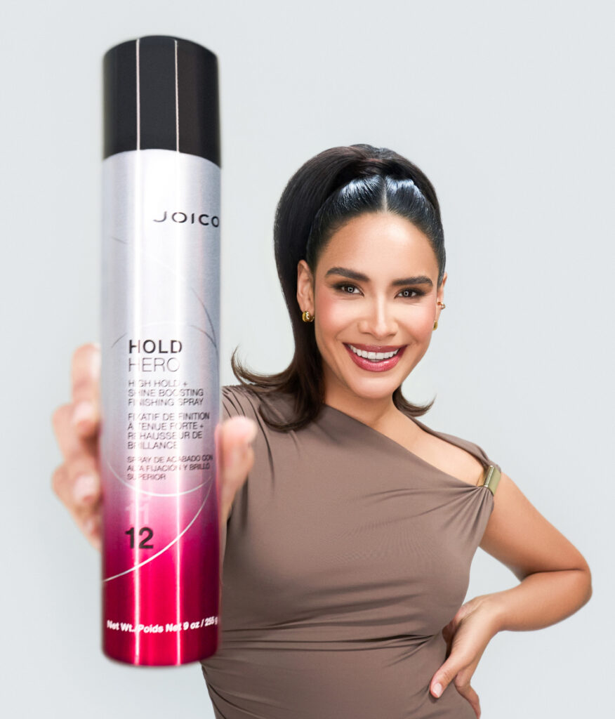 A smiling woman with a high ponytail holds a can of Joico Power Spray Fast-Dry Finishing Spray toward the camera against a plain light background.