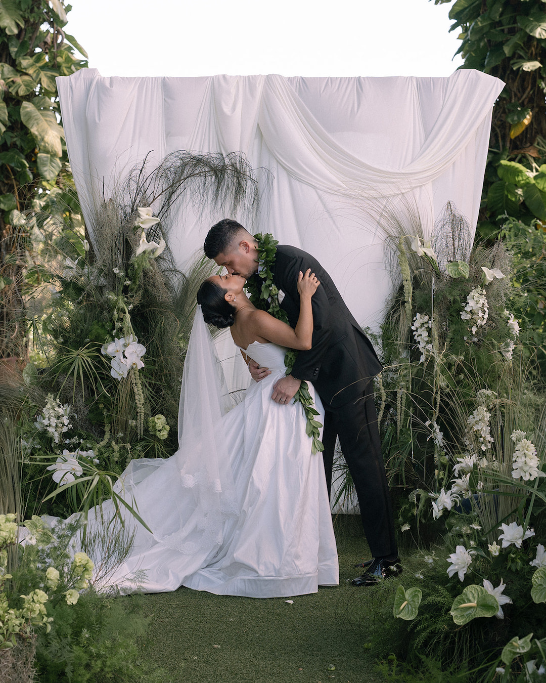 A bride and groom in formal attire kiss in front of a white draped backdrop surrounded by greenery and white flowers at an outdoor wedding.