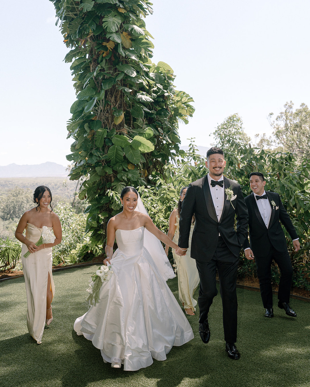 A bride and groom walk hand in hand outdoors, smiling, accompanied by two people in formal attire, surrounded by greenery.