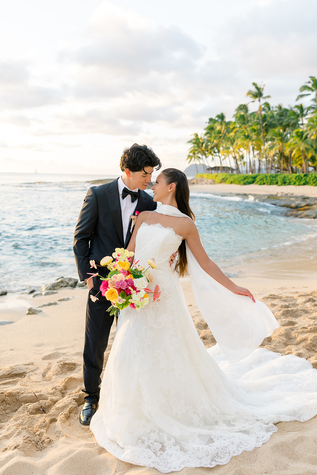 A bride and groom stand on a sandy beach, facing each other, with the ocean and palm trees in the background. The bride holds a bouquet of colorful flowers.
