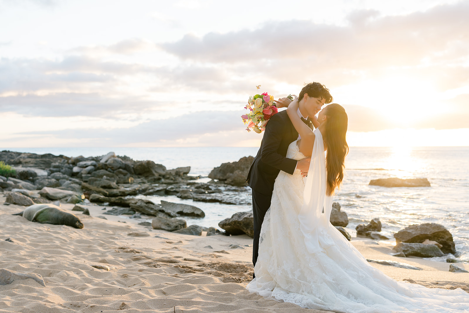 A bride and groom in wedding attire kiss on a sandy beach at sunset, with rocks and the ocean in the background.