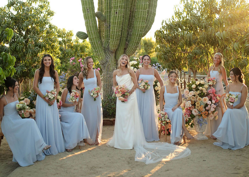 A bride in a white dress stands outdoors with six bridesmaids in light blue dresses, all holding bouquets, near a large cactus and colorful floral arrangements.