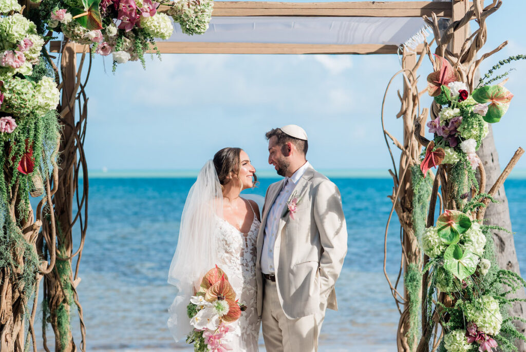 A bride and groom stand under a floral wedding arch on a Islamorada beach, facing each other and smiling, with the ocean in the background.