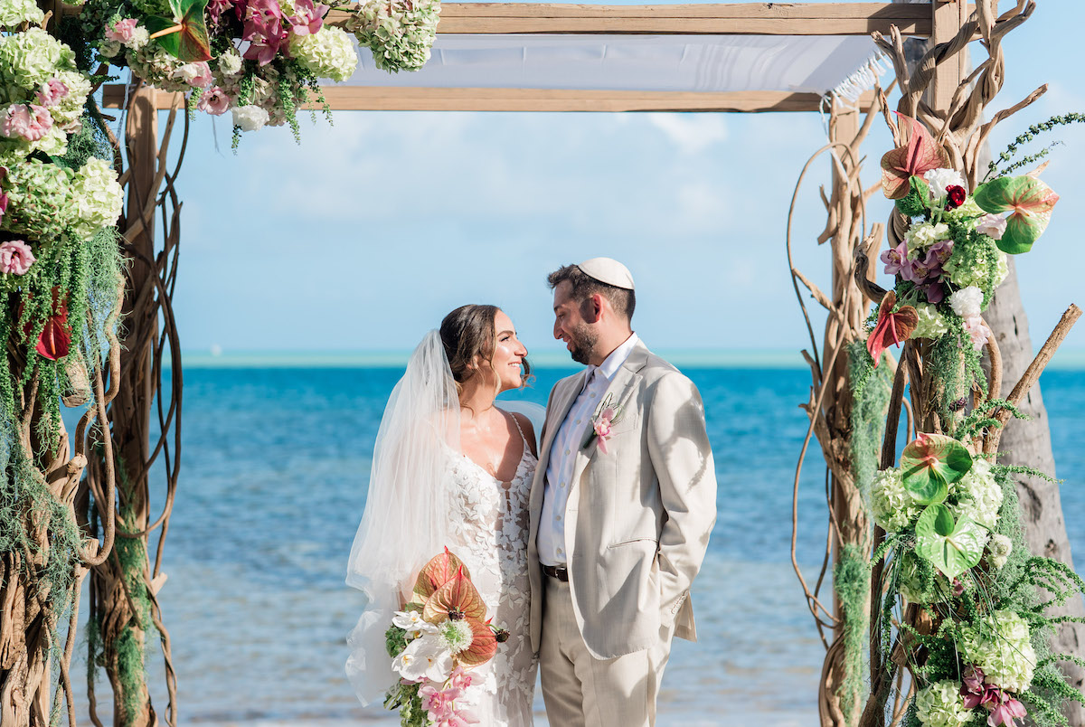 A bride and groom stand under a floral wedding arch on a Islamorada beach, facing each other and smiling, with the ocean in the background.