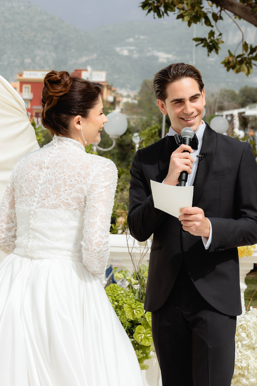 A man in a suit holding a microphone reads from a paper while standing next to a woman in a white lace wedding dress outdoors.