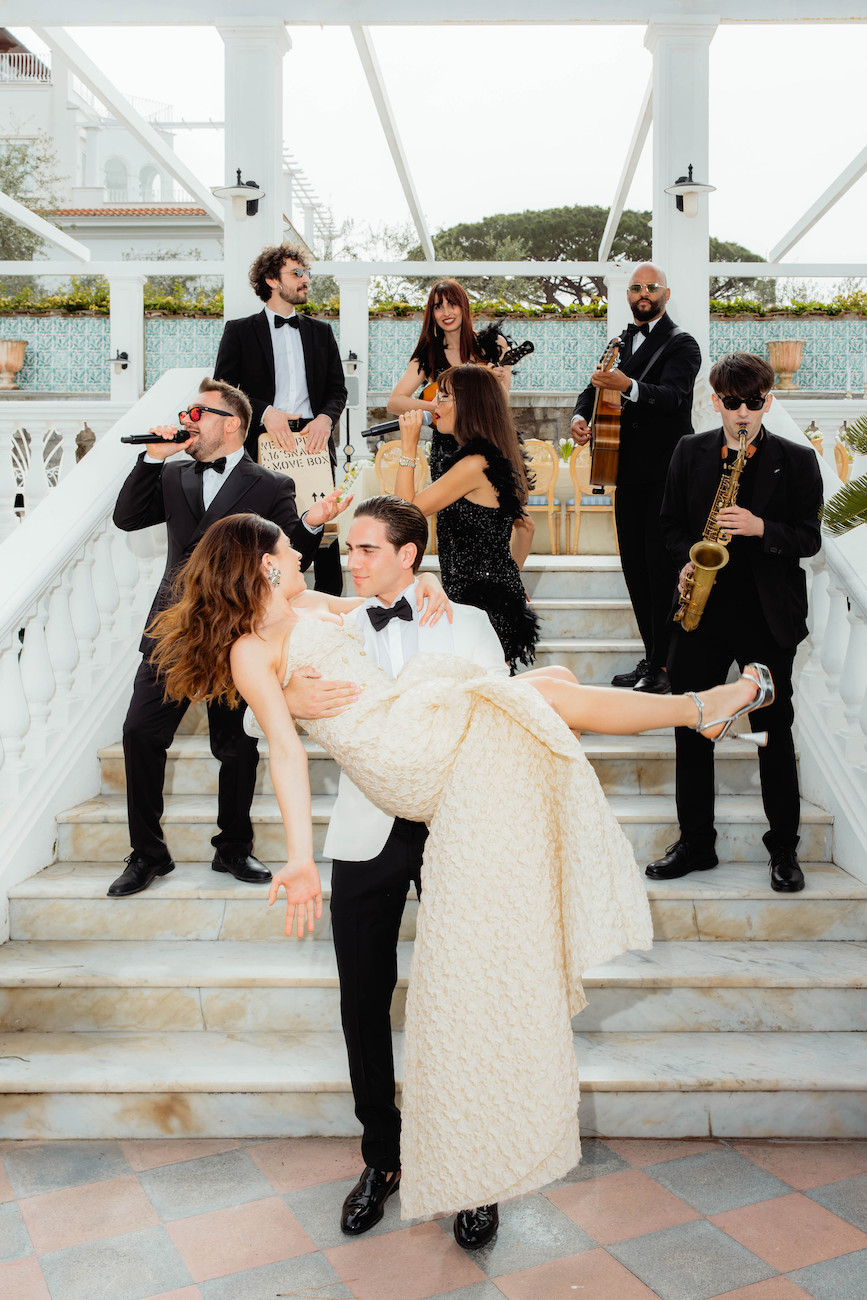 A man in a tuxedo holds a woman in a sparkling gown on a staircase, while a band with various instruments performs behind them.