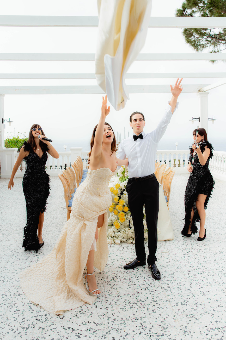A bride throws her bouquet while standing next to a groom, with three guests behind them reaching to catch it at an outdoor wedding.