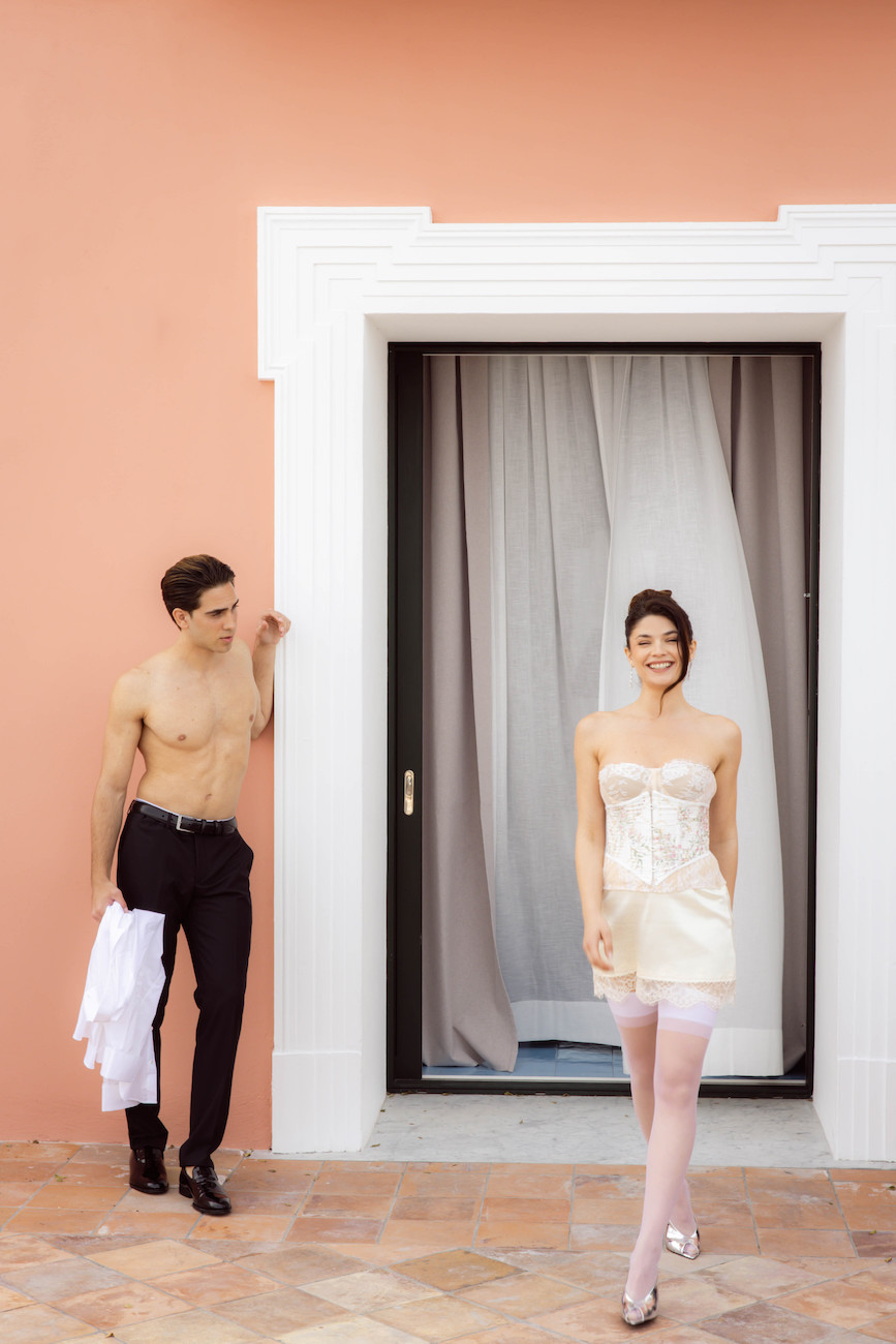 A shirtless man stands by a doorway holding a white shirt, while a woman in a short white dress and pink stockings stands smiling in front of the door against a peach-colored wall.