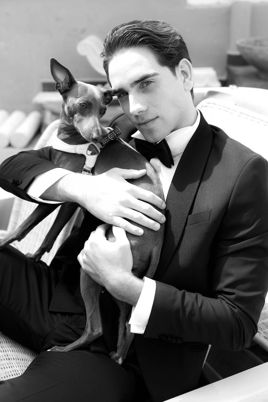A man in a suit sits and holds a small dog in his arms, both looking toward the camera.
