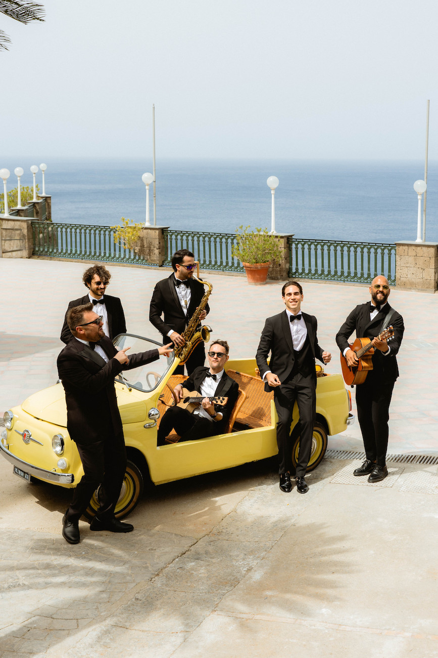 Six men in tuxedos, some holding musical instruments, pose around and inside a small yellow convertible car parked near a seaside promenade.