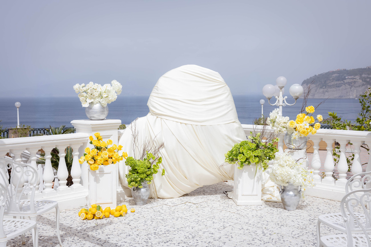 An outdoor terrace overlooking the sea features a large object draped in white fabric, surrounded by white chairs and flower arrangements in yellow, white, and green.