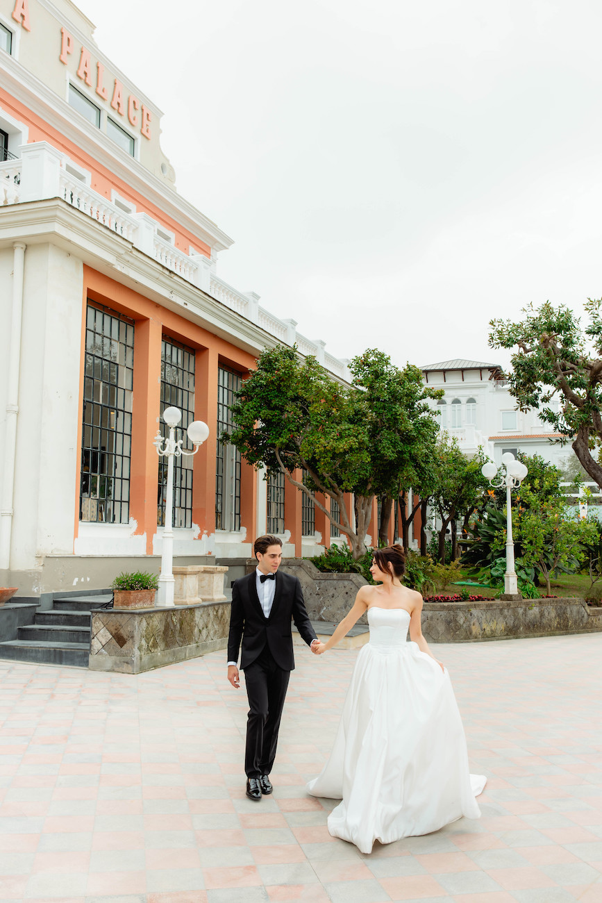 A bride in a white gown and a groom in a black tuxedo walk hand in hand outside a large, elegant building with tall windows and trees nearby.