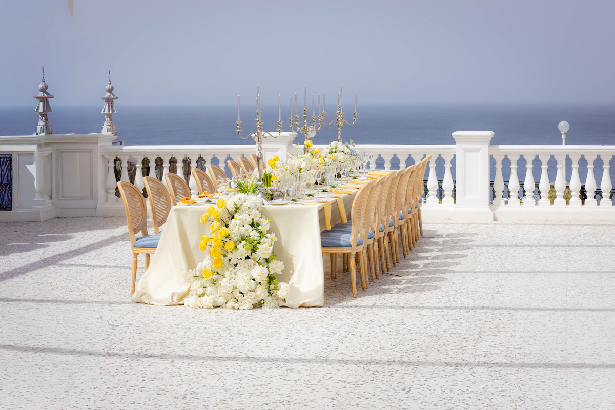 A long table set for a formal outdoor event with yellow and white floral arrangements on a terrace overlooking the sea.