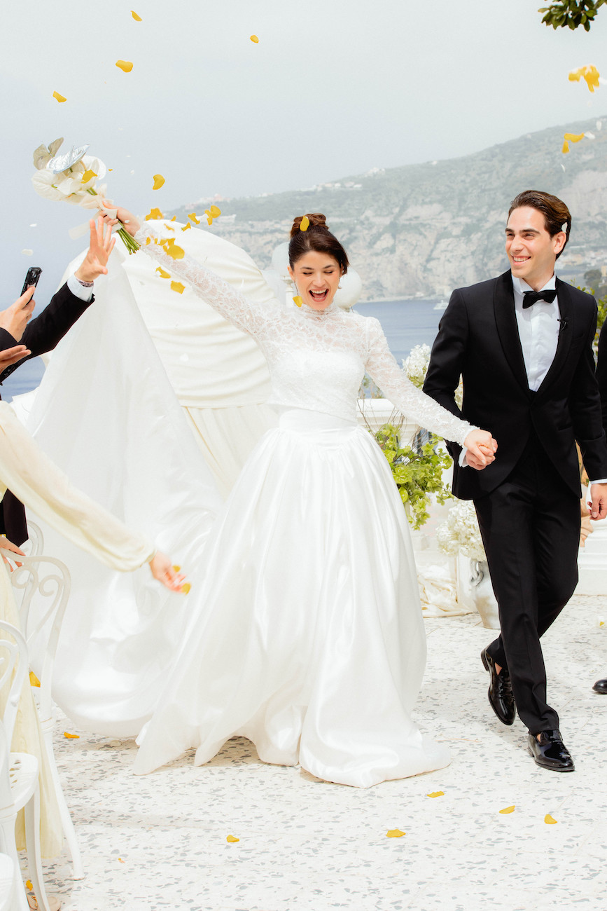 A bride in a white gown and a groom in a black tuxedo walk outside, holding hands and smiling, with flower petals being tossed around them.