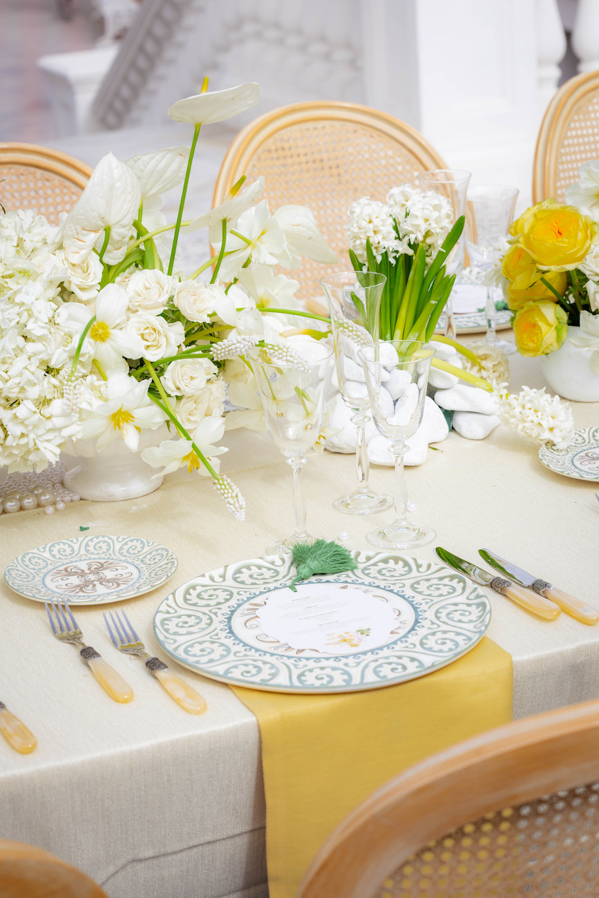 A formal table setting with patterned plates, yellow napkins, cutlery, and floral centerpieces featuring white and yellow flowers.