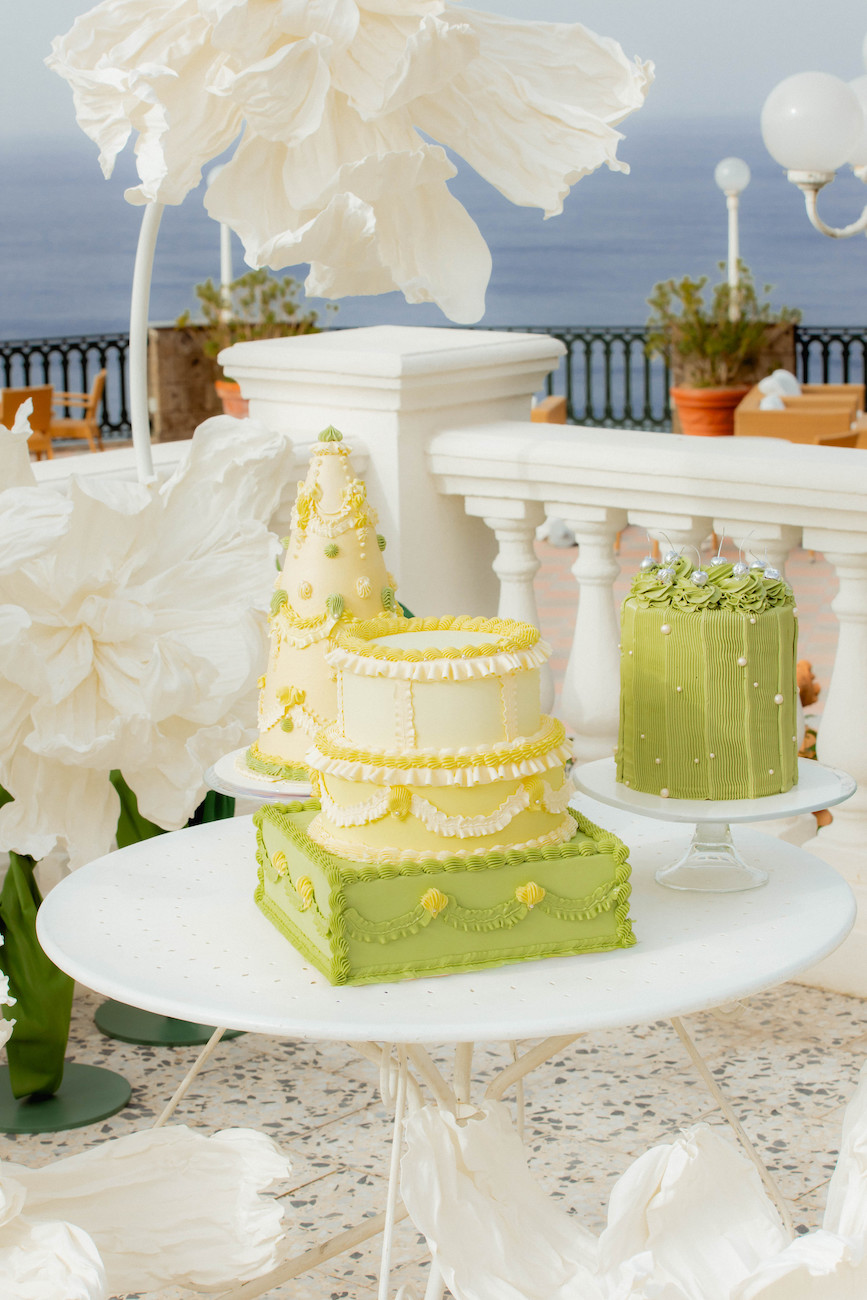 A white table displays three decorative cakes with green and yellow frosting, set outdoors with large white paper flowers and a view of the sea in the background.