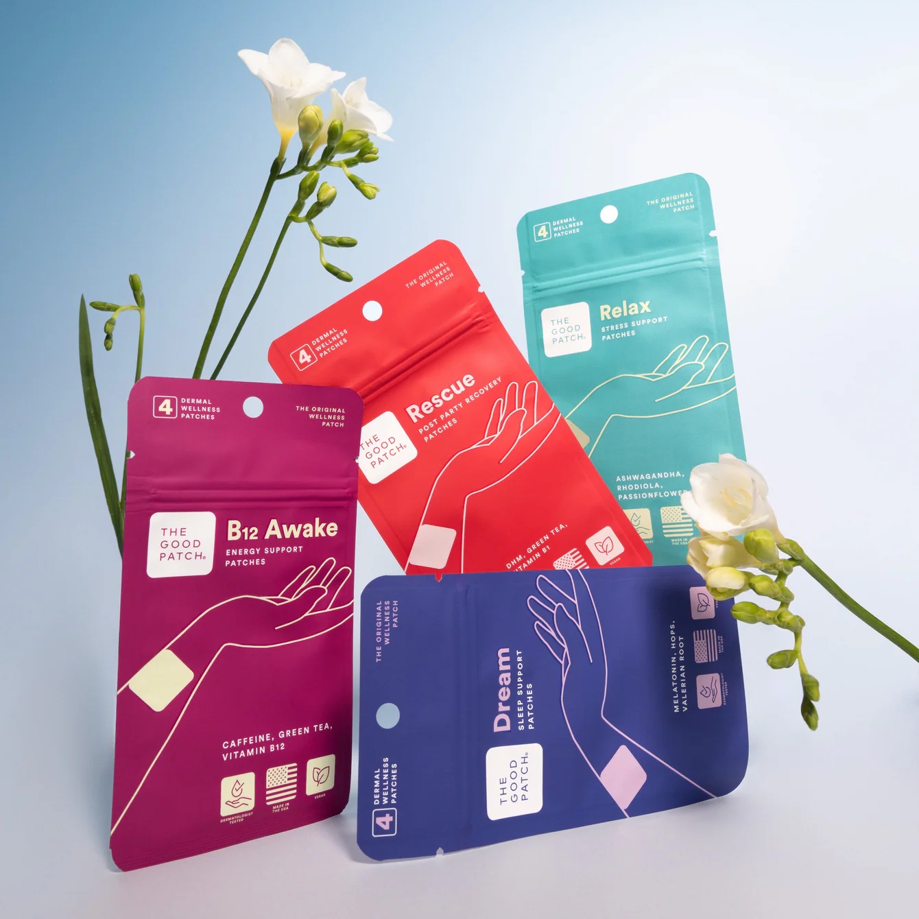 Four colored packets of The Good Patch wellness patches labeled B12 Awake, Rescue, Relax, and Dream, arranged with white flowers against a light blue background.