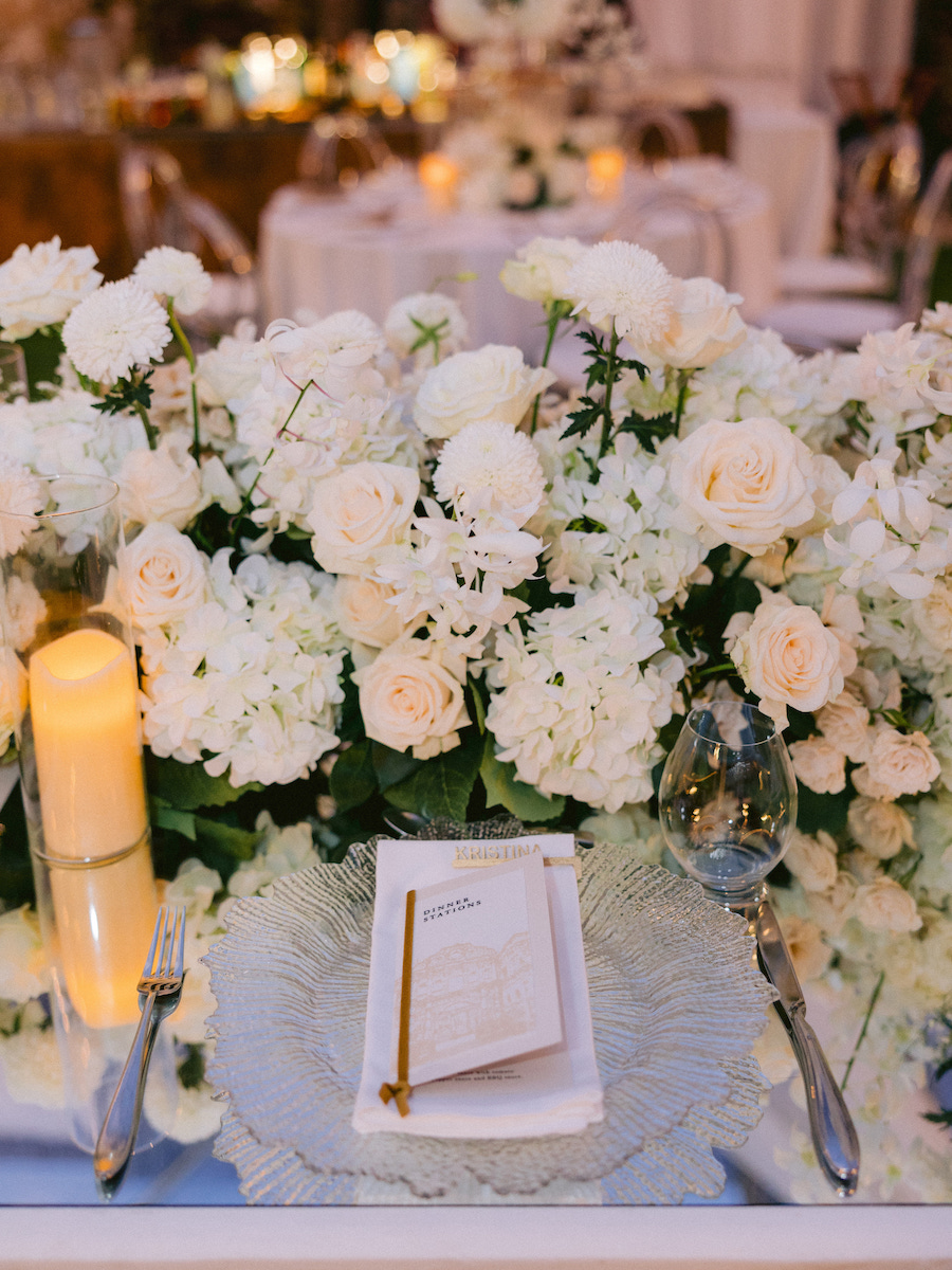 Elegant table setting with a clear glass plate, menu card, gold pen, fork, knife, and wine glass, surrounded by white and pale pink flowers and a lit candle.