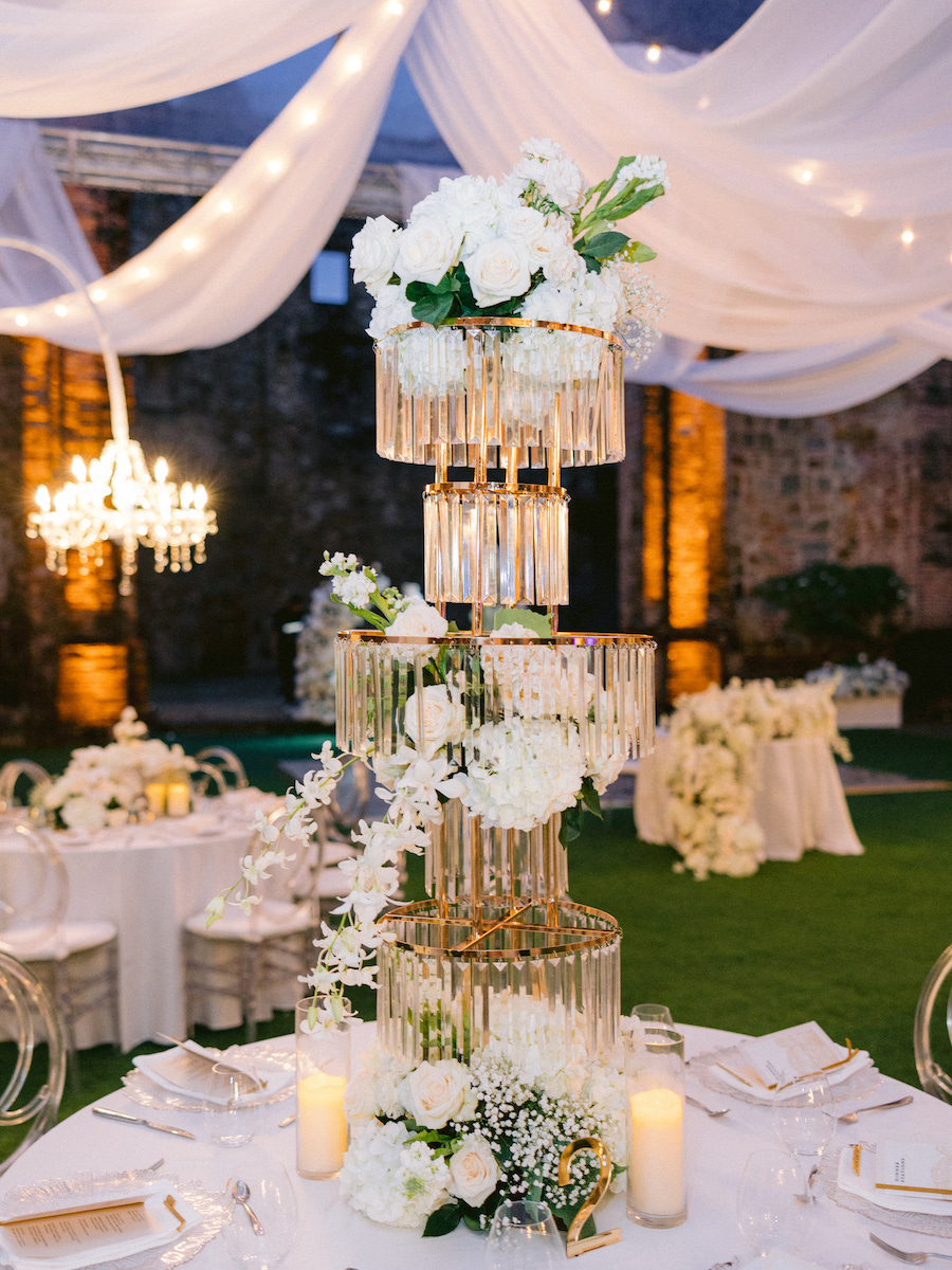 Tall, multi-tiered centerpiece with white flowers and crystal accents on a banquet table, surrounded by candles and elegant wedding decor with draped white fabric overhead.