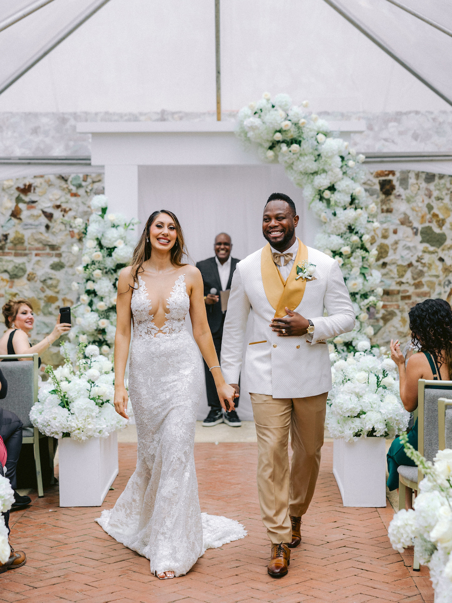 A bride in a white lace gown and a groom in a white jacket with tan pants walk down the aisle together, smiling, with guests seated on either side and white floral decorations behind them.