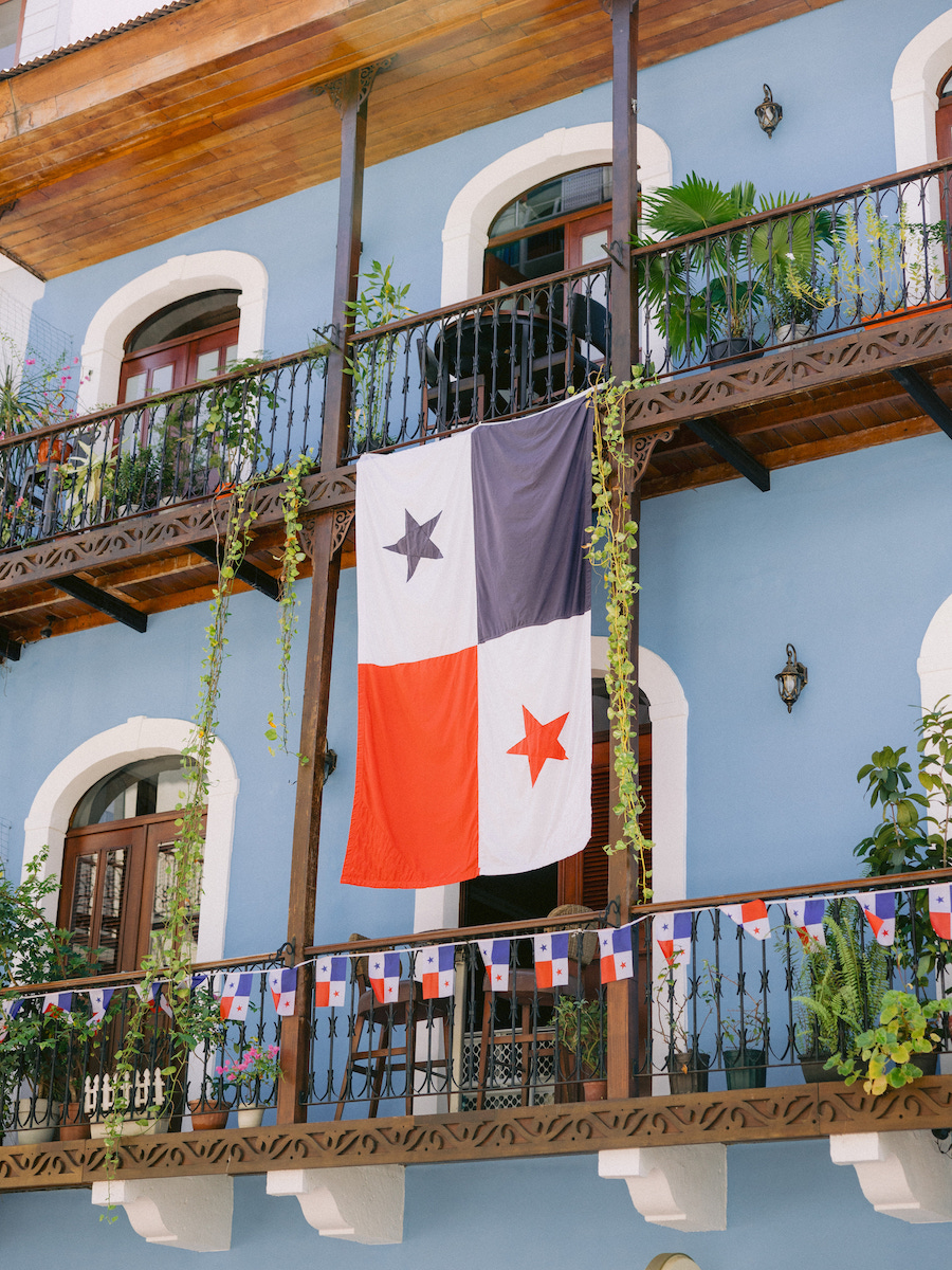 A large Panamanian flag hangs from the wooden balcony of a blue building with plants and decorative flag banners.