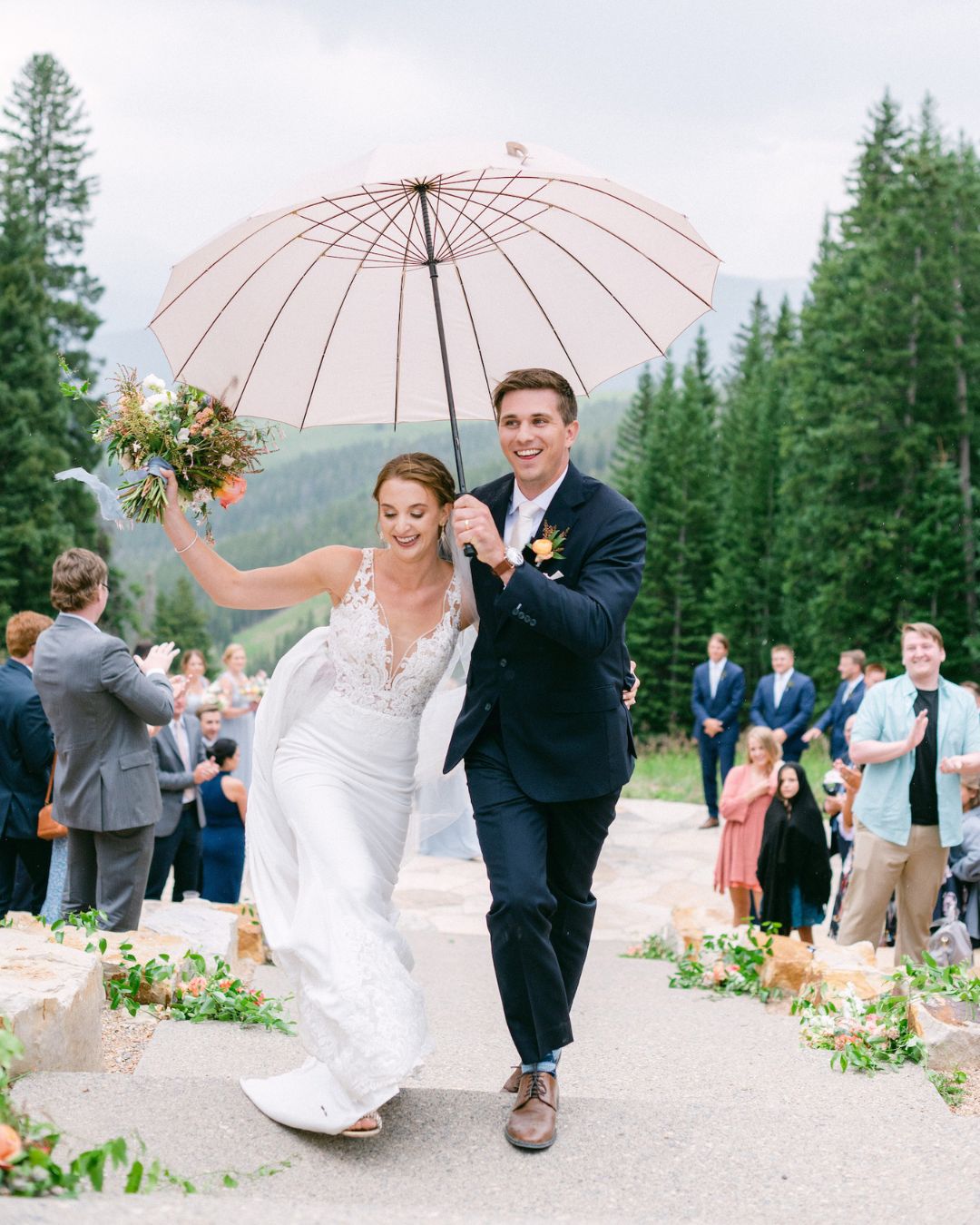 A bride and groom joyfully walk down an outdoor aisle under a pink umbrella, surrounded by guests and pine trees.