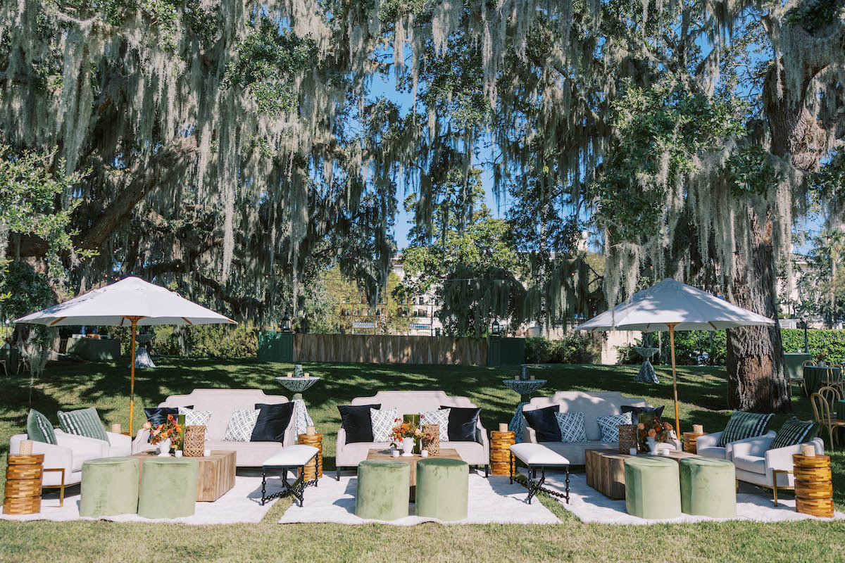Outdoor lounge area with white sofas, green ottomans, umbrellas, and decorative pillows set under large trees with hanging moss, on a sunny day.
