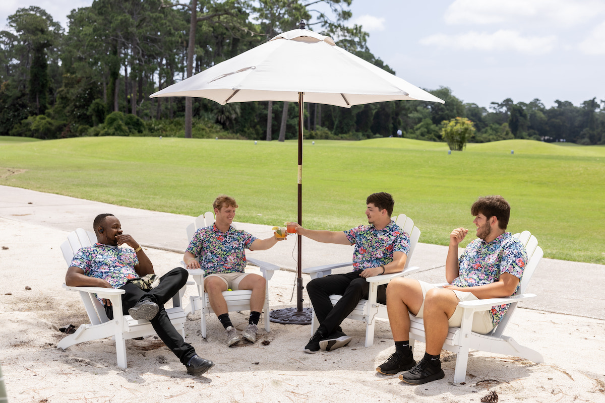 Four men in matching shirts sit in white chairs under a large umbrella on a sandy area, with a green golf course and trees in the background. Two of them are clinking drinks together.