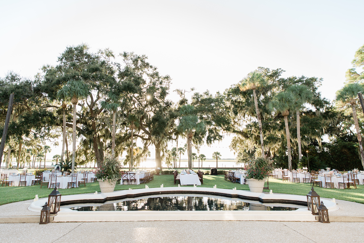 Outdoor event setup with tables and chairs arranged around a rectangular reflecting pool, surrounded by grass and large trees under a bright sky.