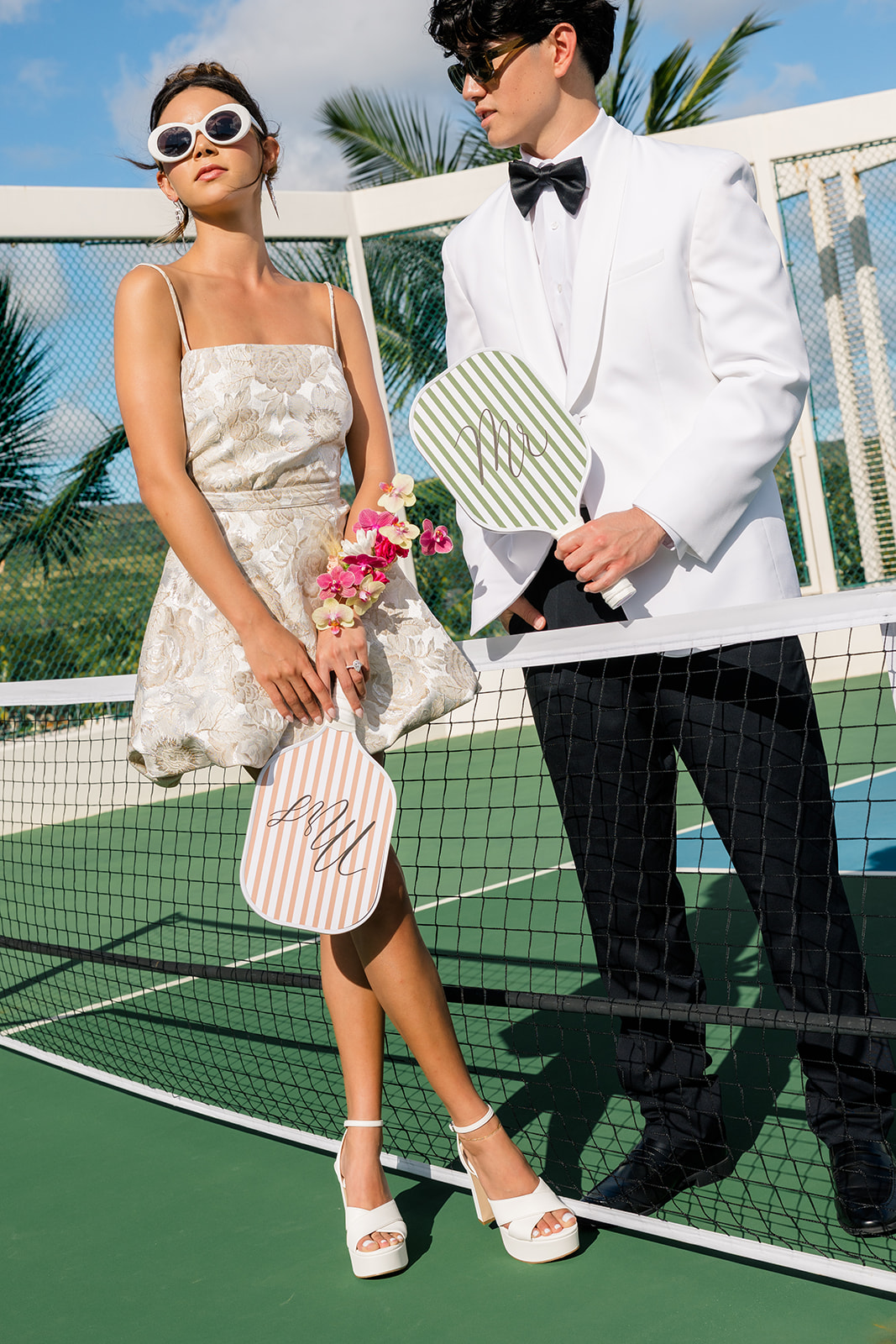 Two people in formal attire stand on a tennis court holding paddles, with a net between them and palm trees in the background.