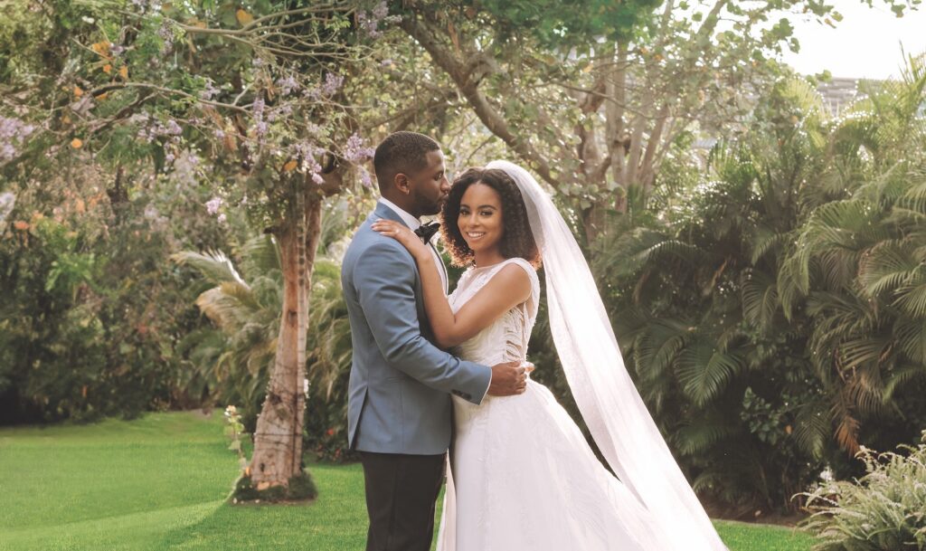 Bride and groom pose together outdoors; the groom wears a blue suit, and the bride wears a white dress with a long veil. Lush greenery and trees appear in the background.
