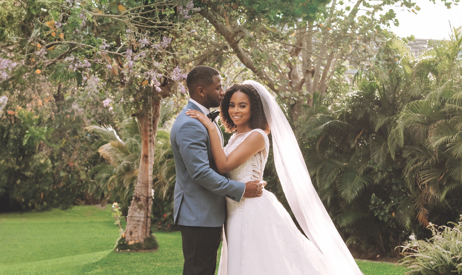 Bride and groom pose together outdoors; the groom wears a blue suit, and the bride wears a white dress with a long veil. Lush greenery and trees appear in the background.