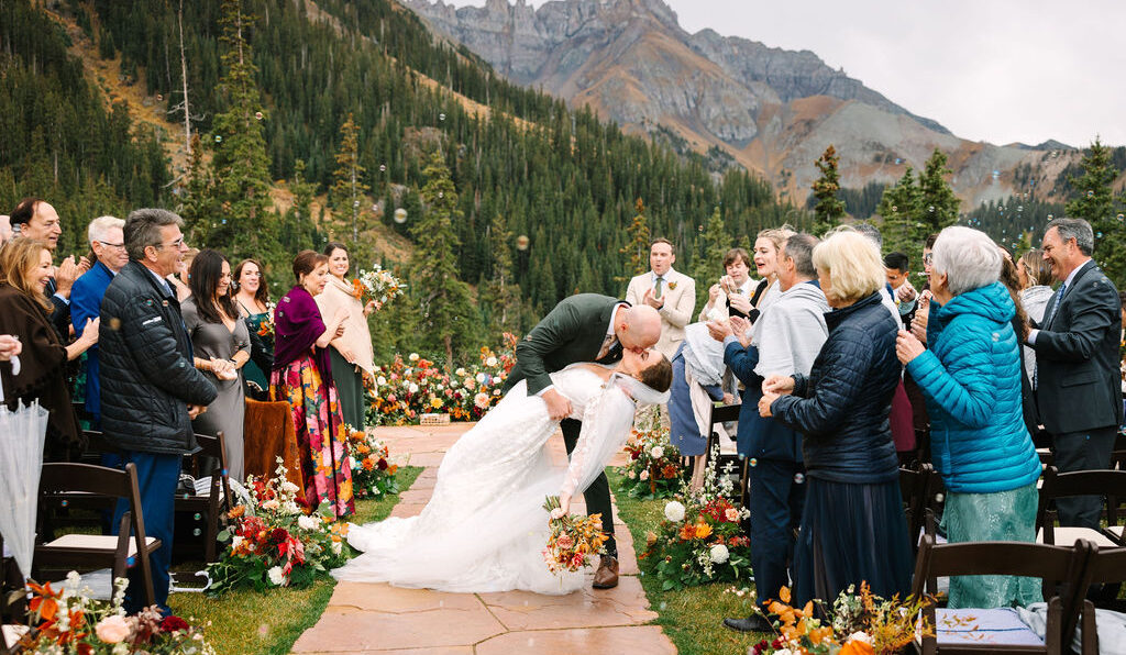 A bride and groom kiss at an outdoor mountain wedding ceremony, surrounded by guests and autumn floral arrangements on a stone path.