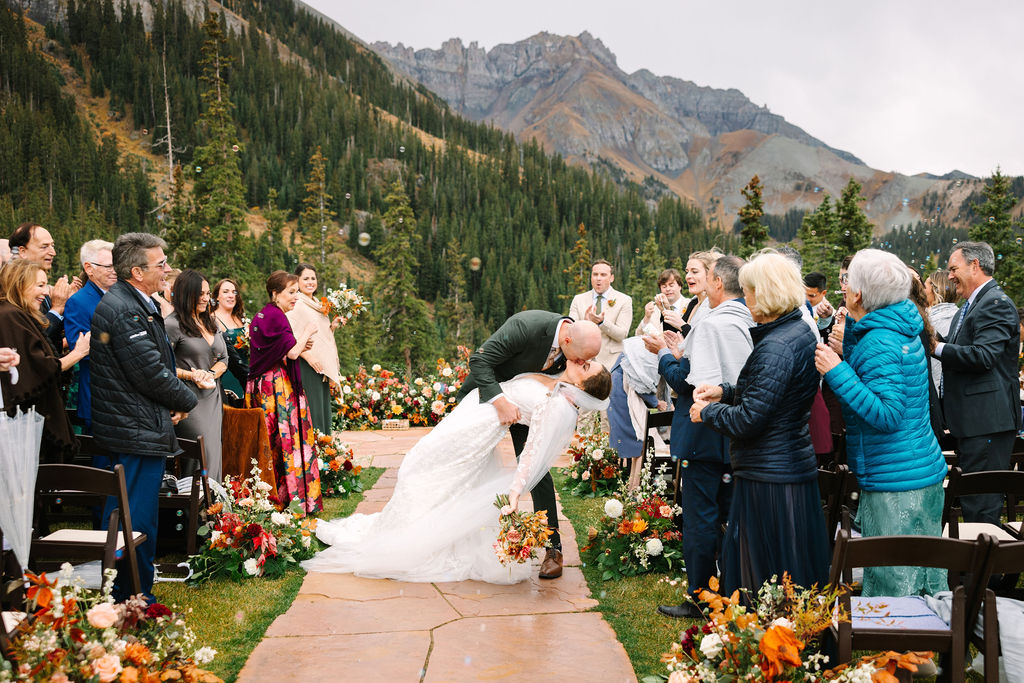 A bride and groom kiss at an outdoor mountain wedding ceremony, surrounded by guests and autumn floral arrangements on a stone path.