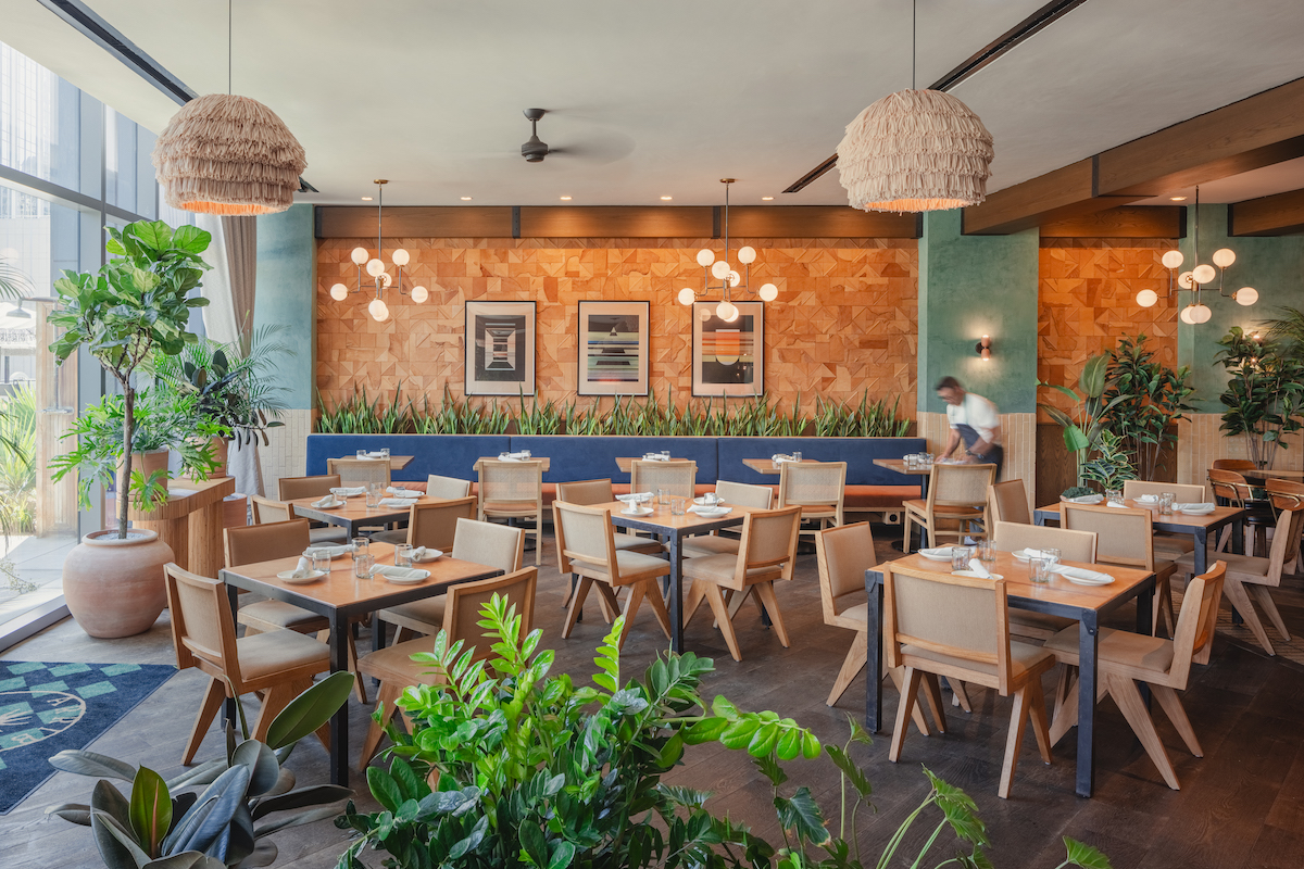 Modern restaurant interior with wooden tables and chairs, green plants, pendant lights, and a person setting a table near a warm-toned wall with framed artwork.