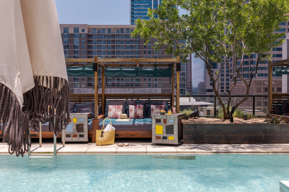 Daybeds with cushions and throw pillows are placed beside a clear outdoor pool, with city buildings and trees visible in the background.