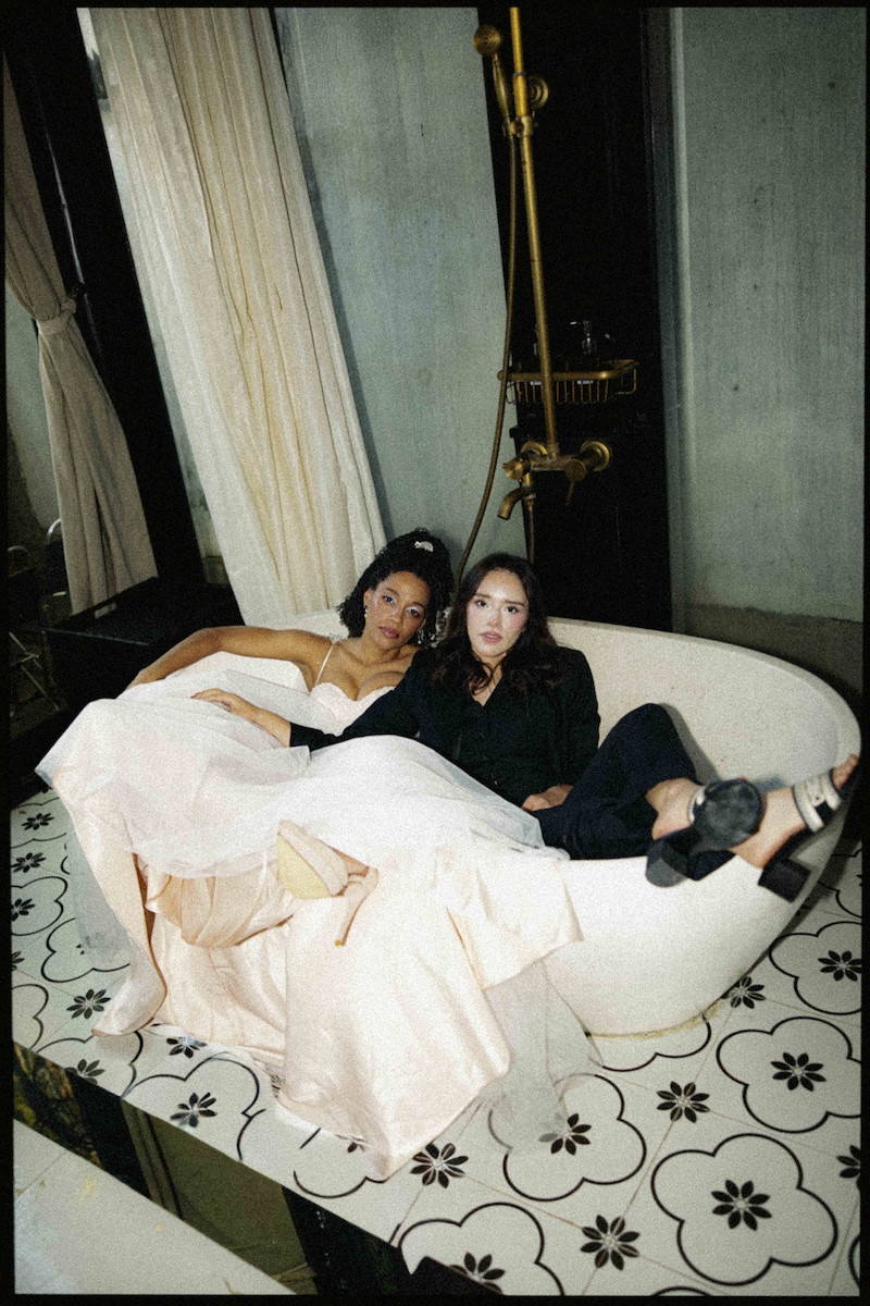 Two women sit together in a white freestanding bathtub on a patterned tile floor; one wears a formal dress, the other casual clothes.