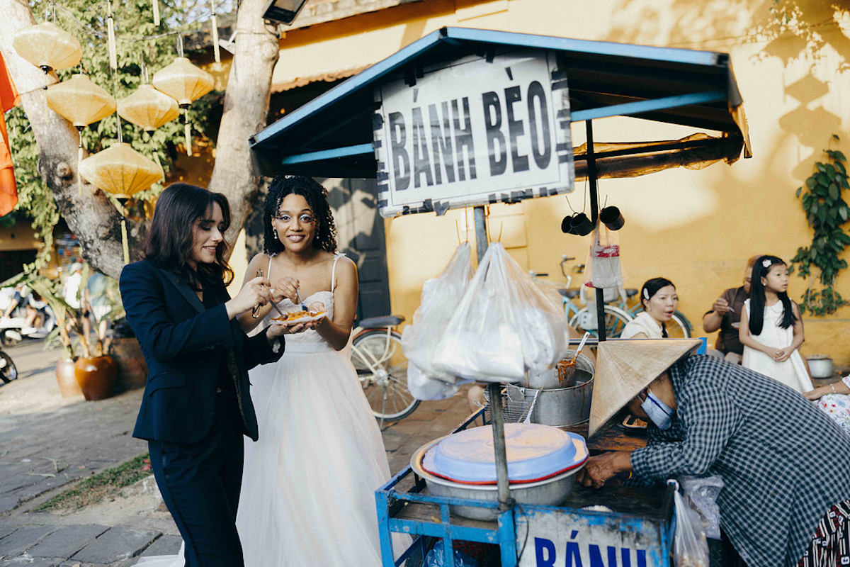 Two women, one in a suit and one in a white dress, eat food at a Bánh Bèo street food stall while a vendor in a conical hat serves them.