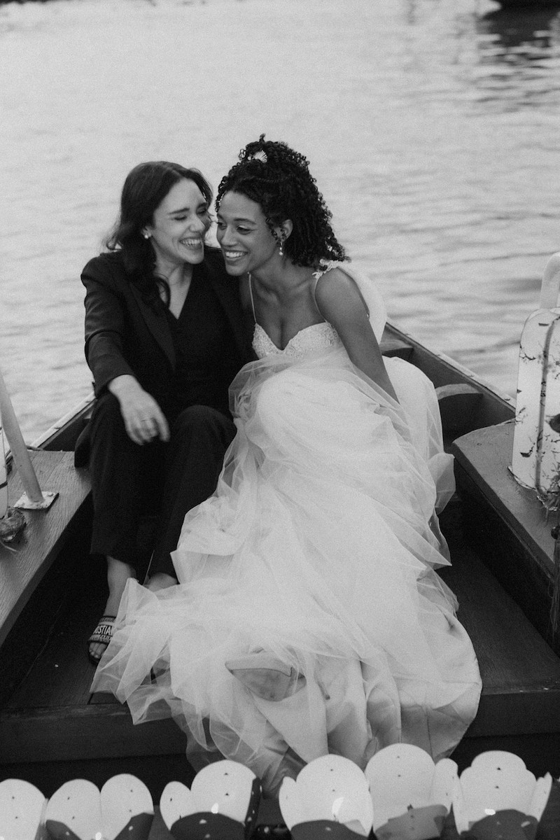Two women sit closely together on a small boat; one wears a suit, and the other wears a wedding dress. The water is visible in the background.