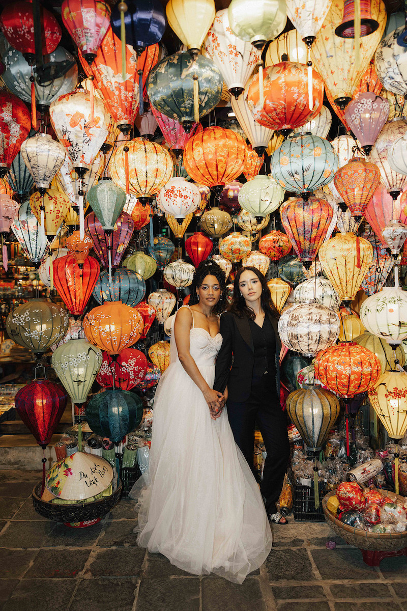 Two women, one in a white wedding dress and one in a dark suit, stand side by side in front of a colorful display of hanging lanterns.