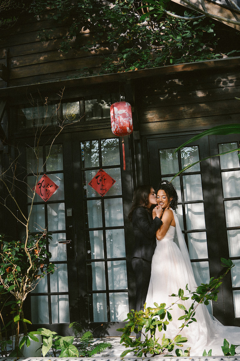 Two women in wedding attire embrace and smile in front of a building with glass doors, red lantern, and Chinese New Year decorations, surrounded by greenery and sunlight.