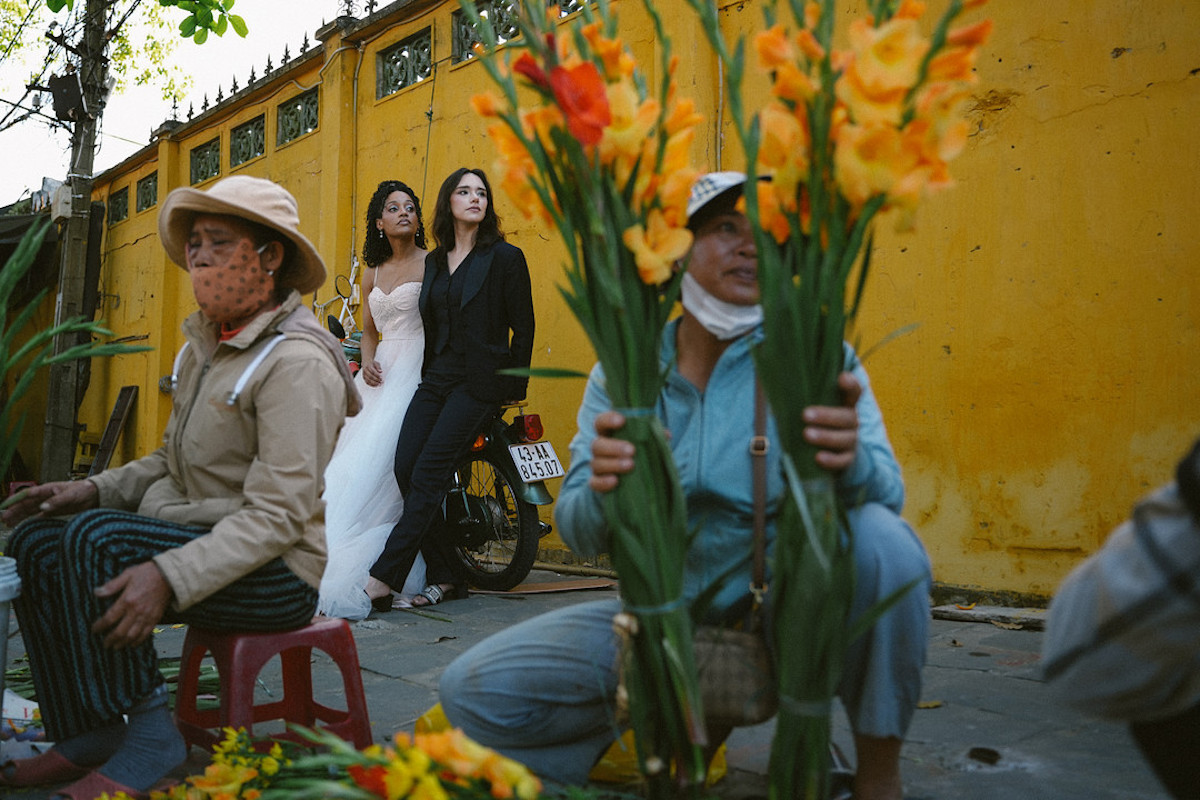 Two flower vendors sit by a yellow wall, while two women in formal dresses walk past in the background. Bright orange flowers are in the foreground.