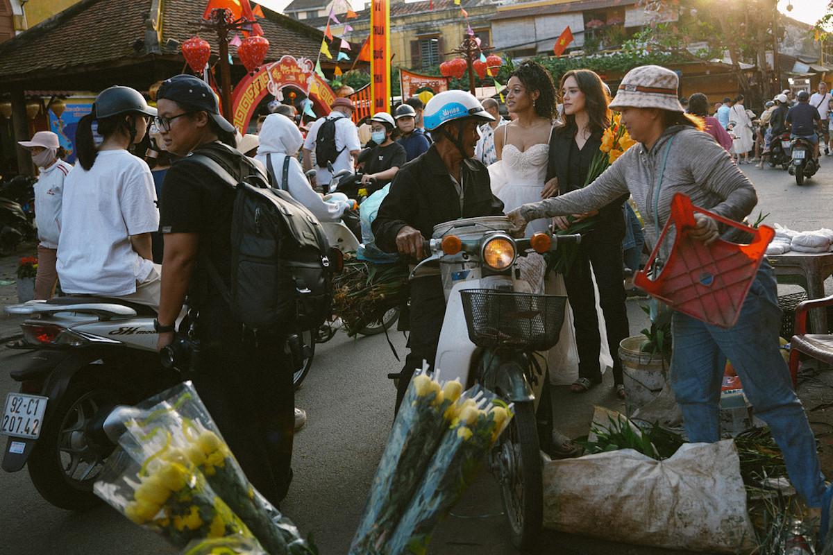 A busy street market scene with people, motorbikes, flower vendors, and a woman in a white dress standing among the crowd.