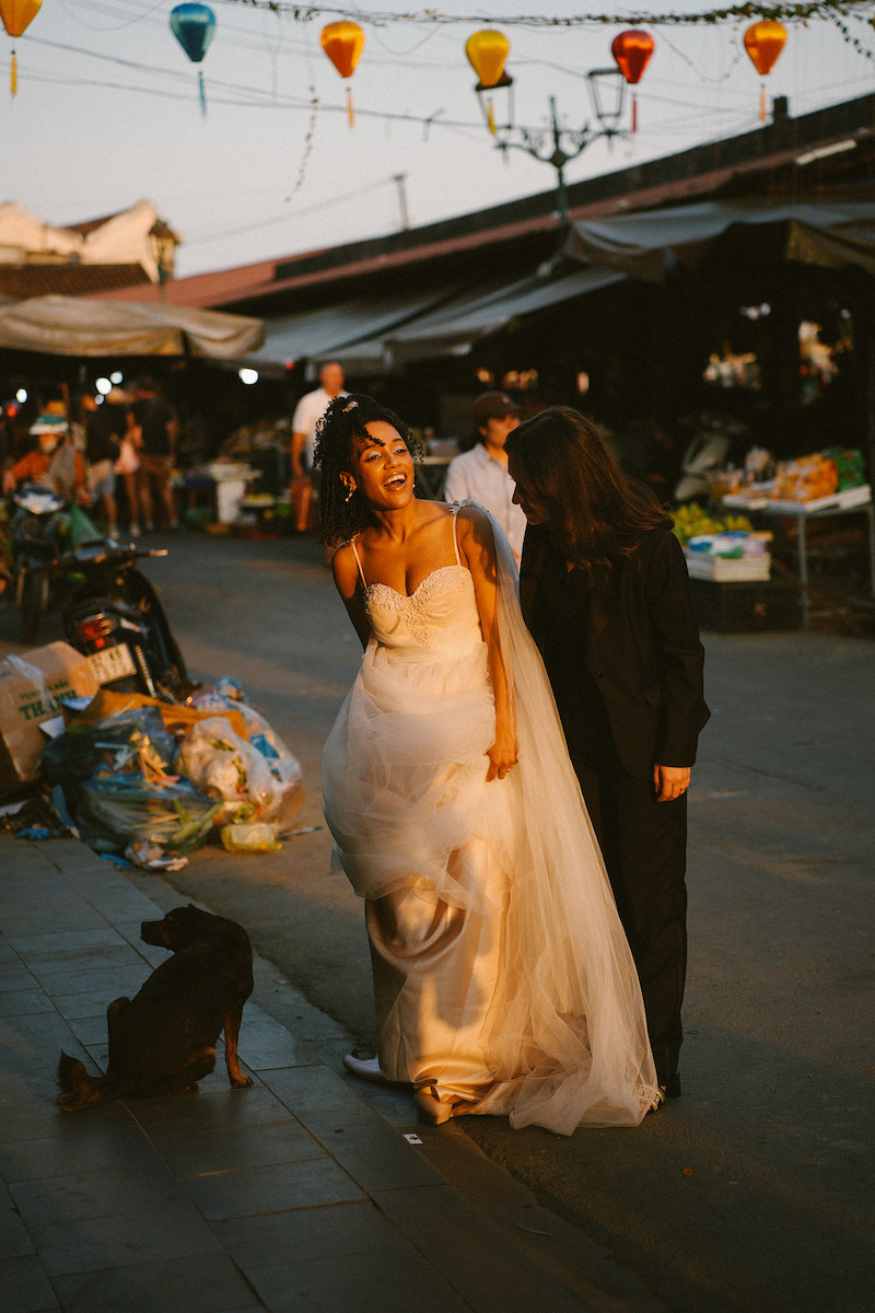 A bride and another person walk together in an outdoor market at sunset, with a dog sitting nearby and stalls in the background.