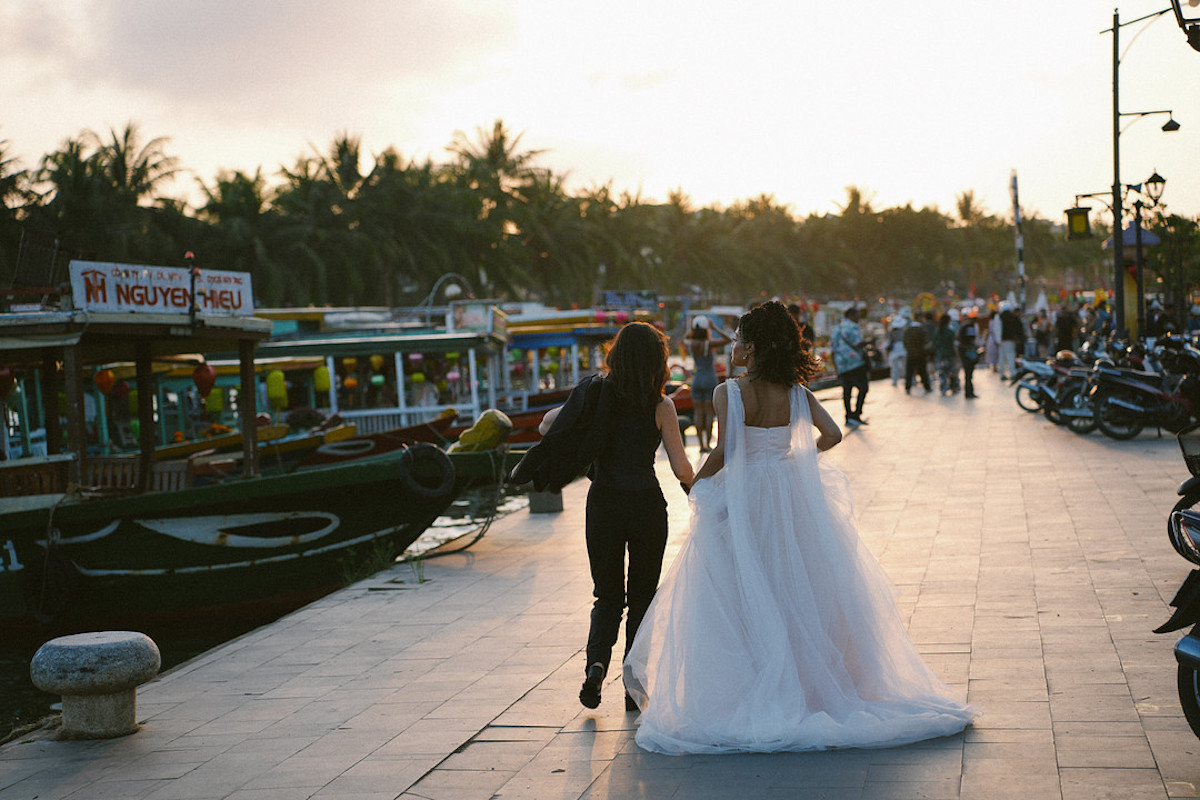 A bride in a white dress walks beside another woman along a riverside promenade lined with boats, people, and parked motorcycles at sunset.