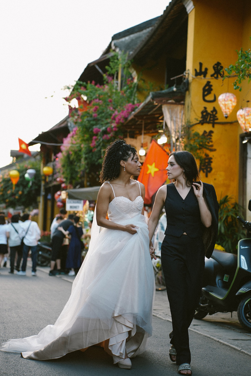 Two women walk hand in hand down a street; one wears a white wedding dress, the other a black suit. They are surrounded by lanterns, flowers, and colorful buildings.