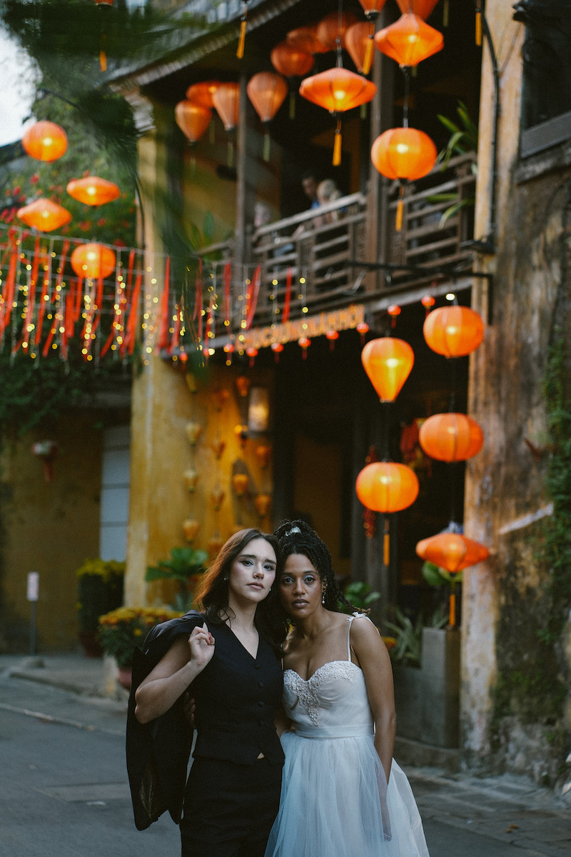 Two women pose together on a street decorated with hanging orange lanterns, one in a black outfit and the other in a white dress, in front of a yellow building.