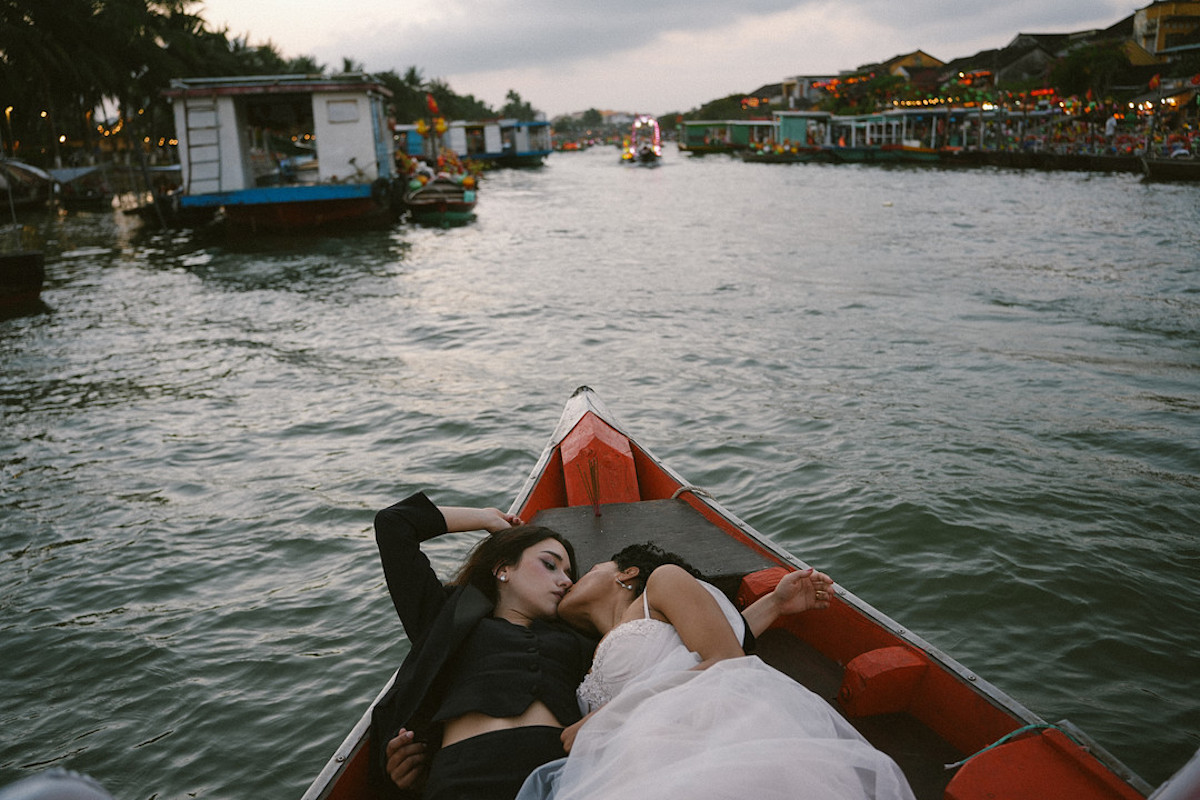 Two people lie close together at the front of a boat, floating on a river with buildings and boats lining the shore under a cloudy sky.