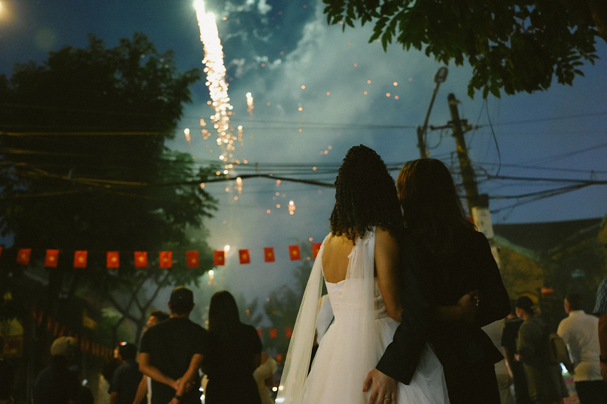 A couple in wedding attire stands close together, watching fireworks at night with a crowd. Red lanterns and trees are visible in the background.